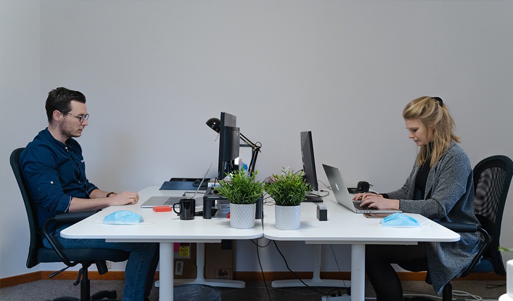 Two people working opposite one another at a white desk.
