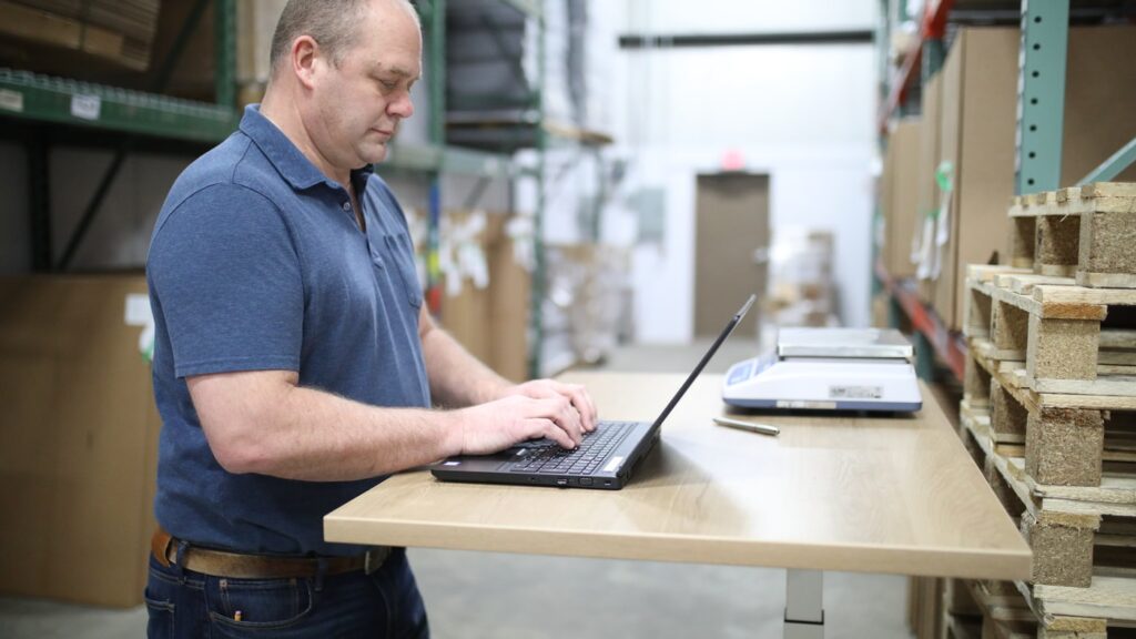 A man in a warehouse typing on a laptop.